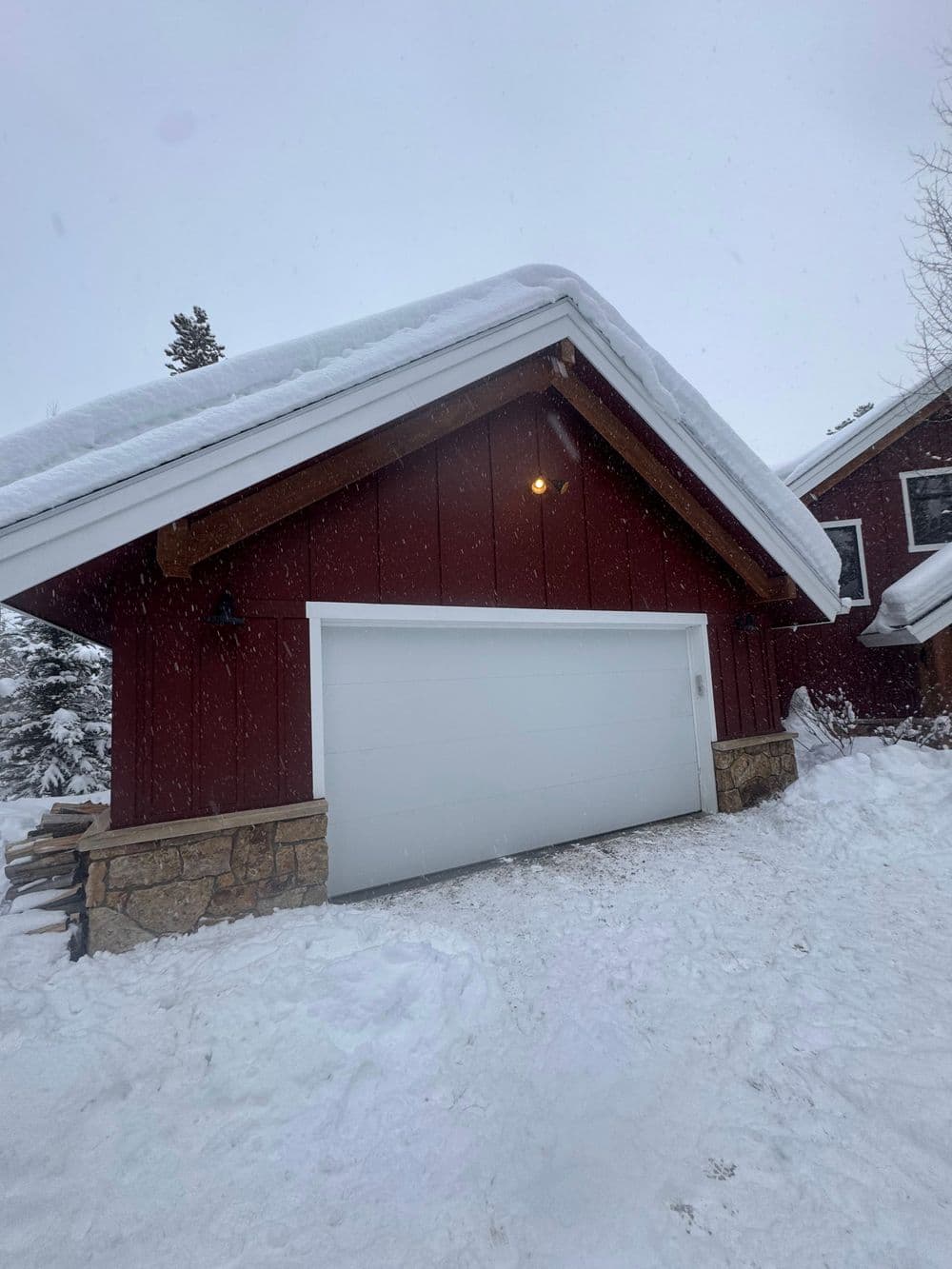Red house with a stone foundation, snow-covered roof, and snowy driveway in winter scenery.