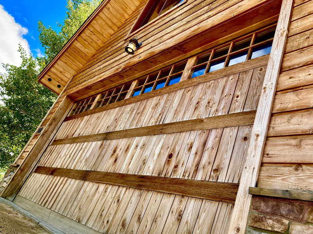 Wooden garage door with rustic design, framed by a log cabin exterior and blue sky above.
