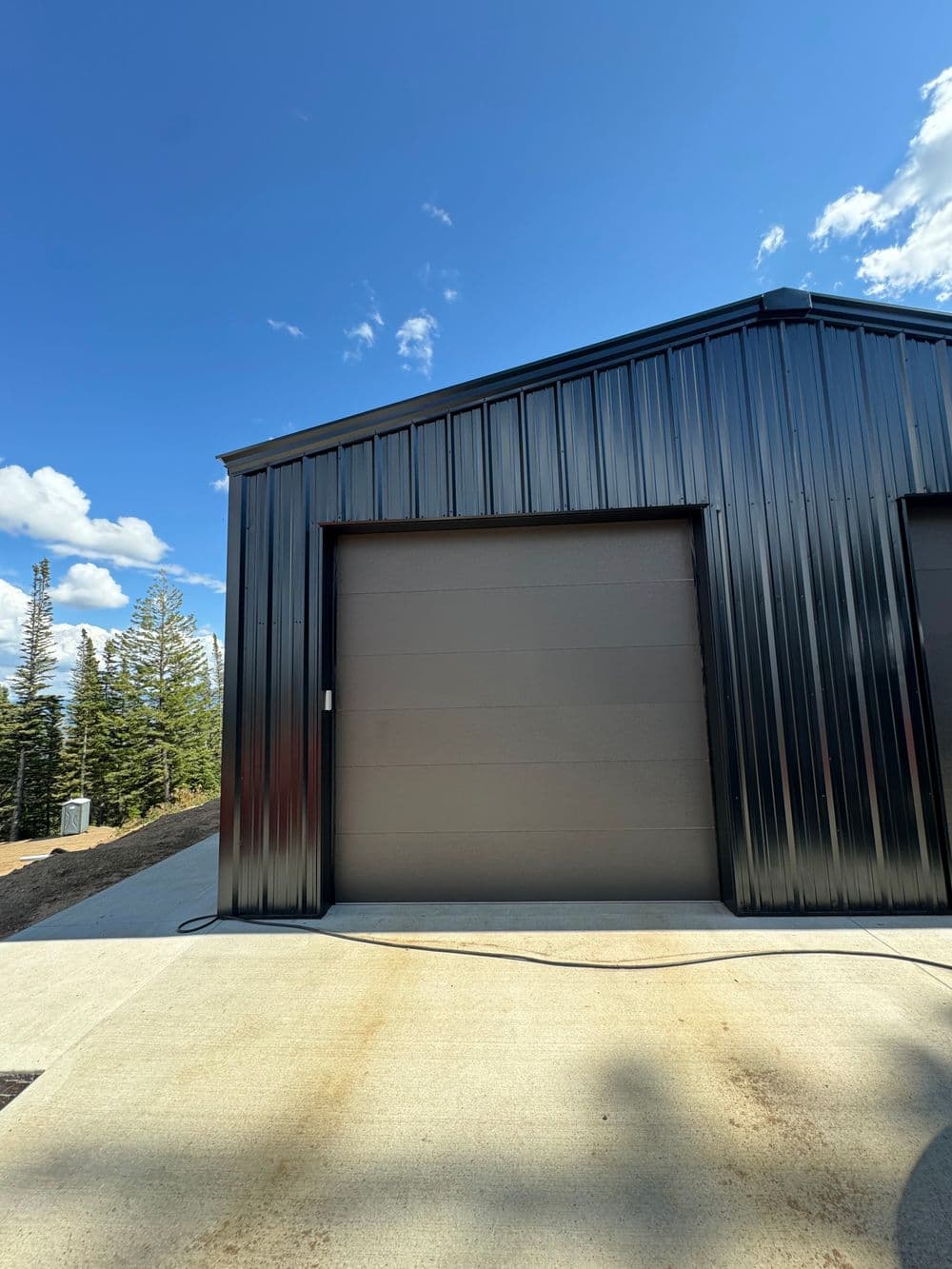 Modern black metal garage with large doors against a blue sky and pine trees.