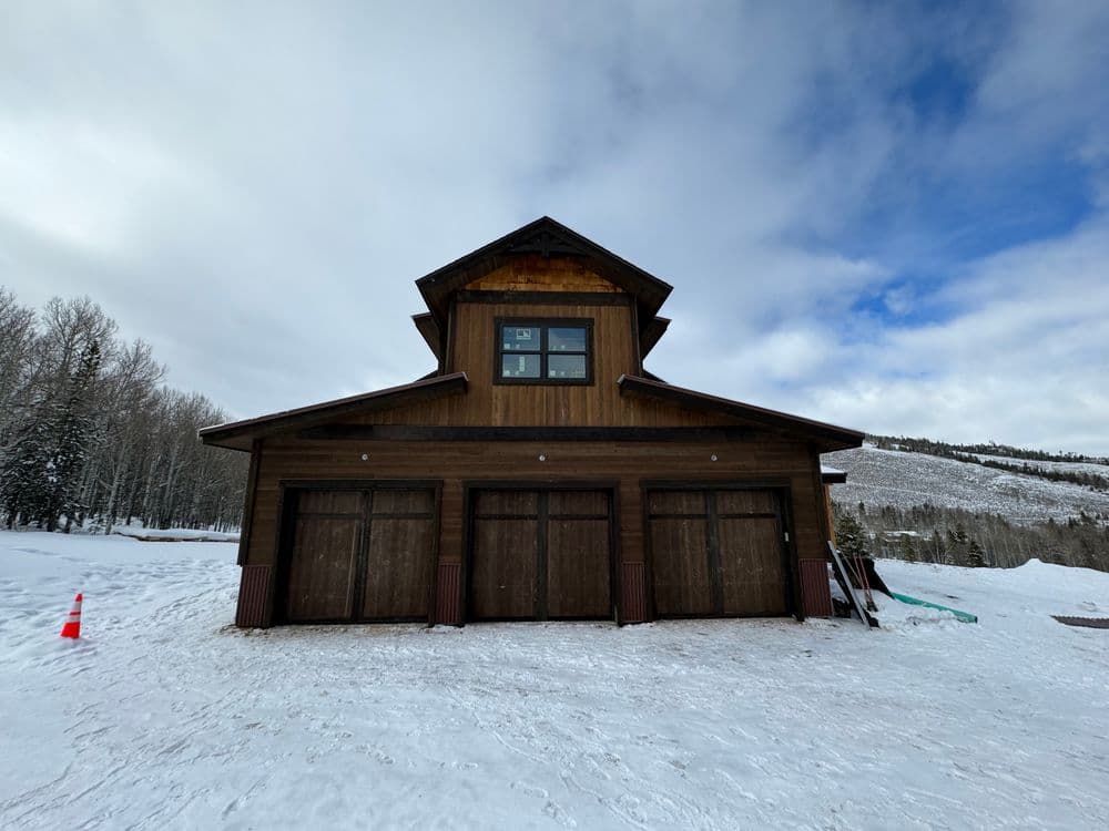 Snow-covered landscape featuring a modern wooden garage with three doors and mountain backdrop.