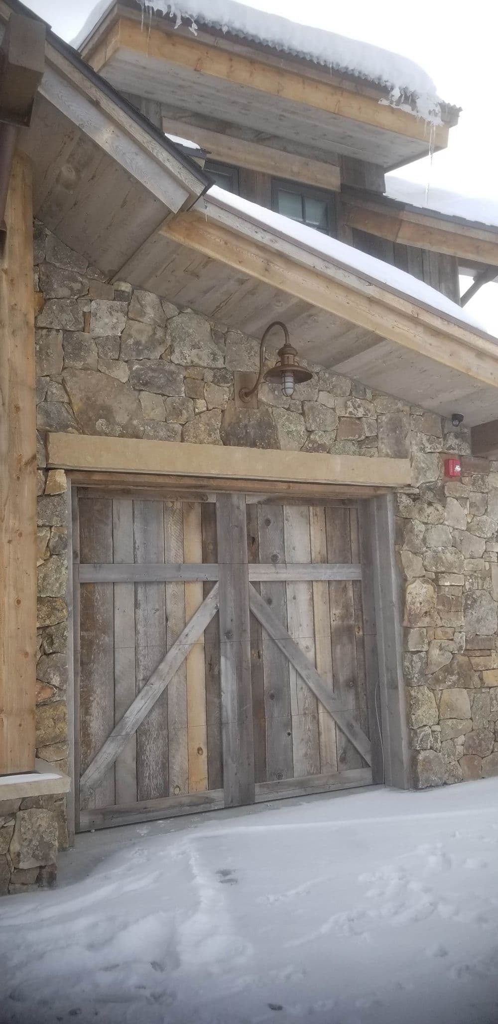 Rustic barn door with stone wall, wooden details, and snow-covered roof in a mountain setting.