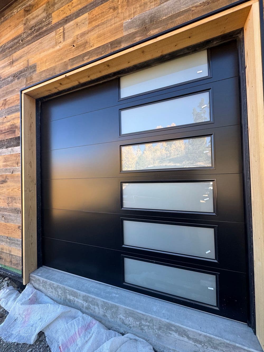 Modern black garage door with glass panels, framed by rustic wooden exterior.