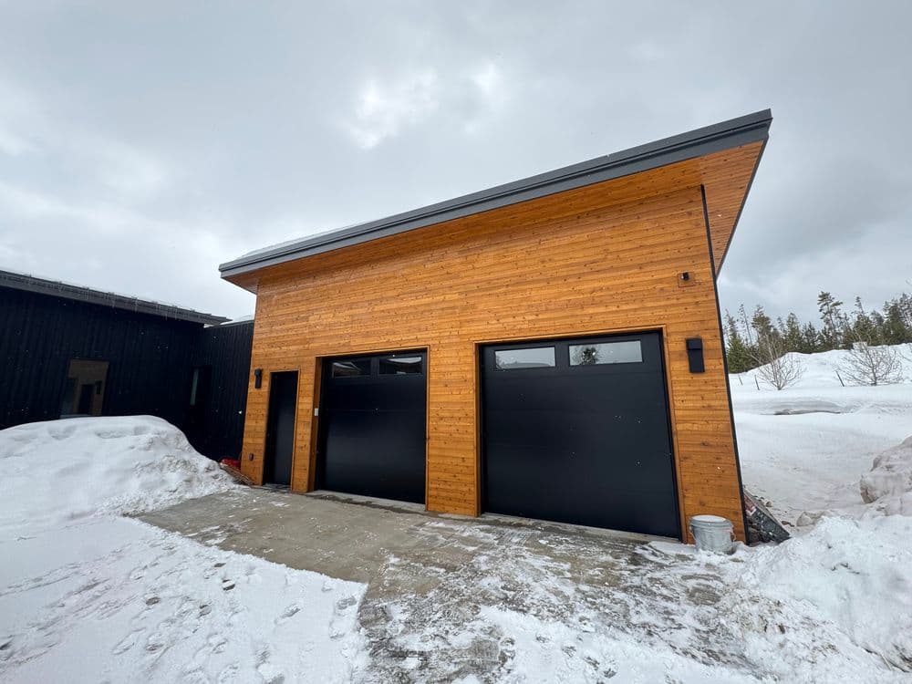 Modern wooden garage with black doors surrounded by snow on a cloudy day.