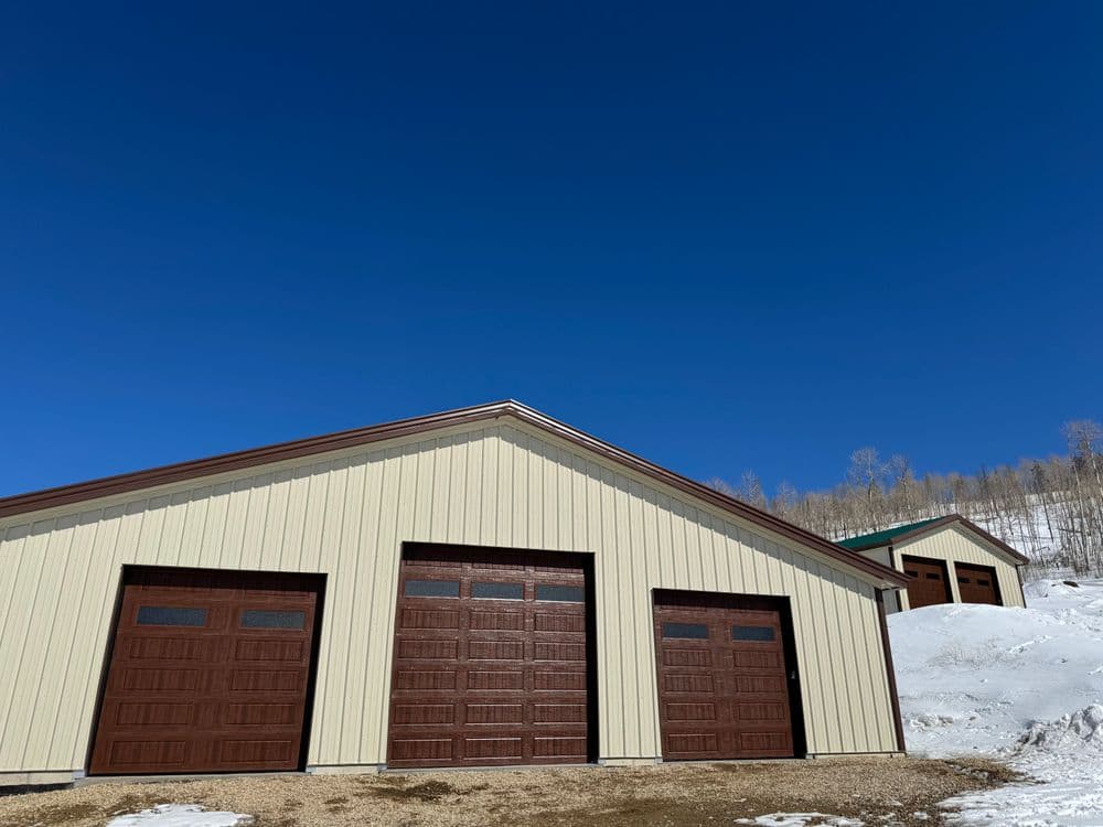 Snow-covered landscape with a large metal warehouse featuring three brown garage doors under a clear blue sky.