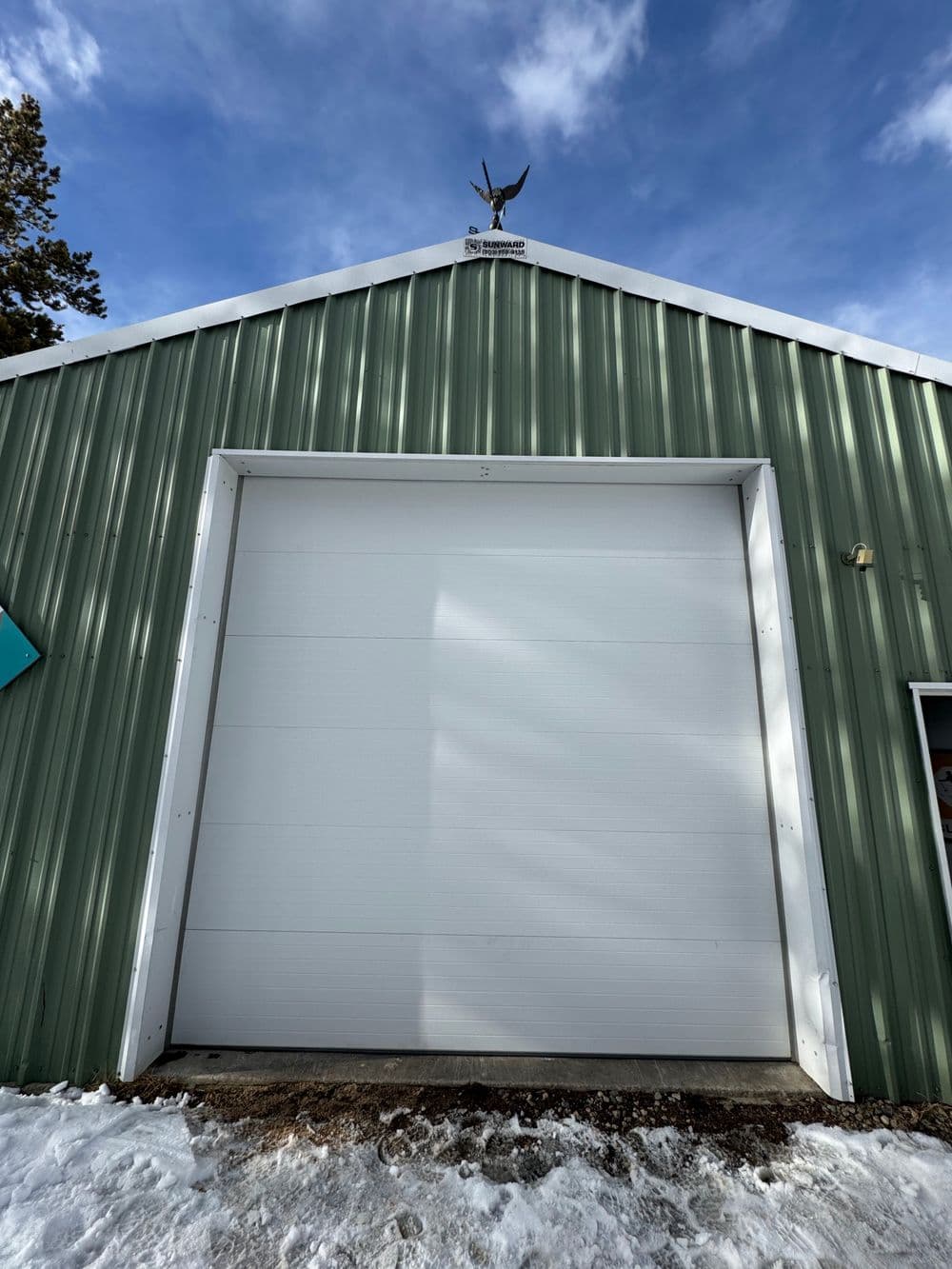 Green metal garage with a white door and eagle decoration against a blue sky. Snow on the ground.