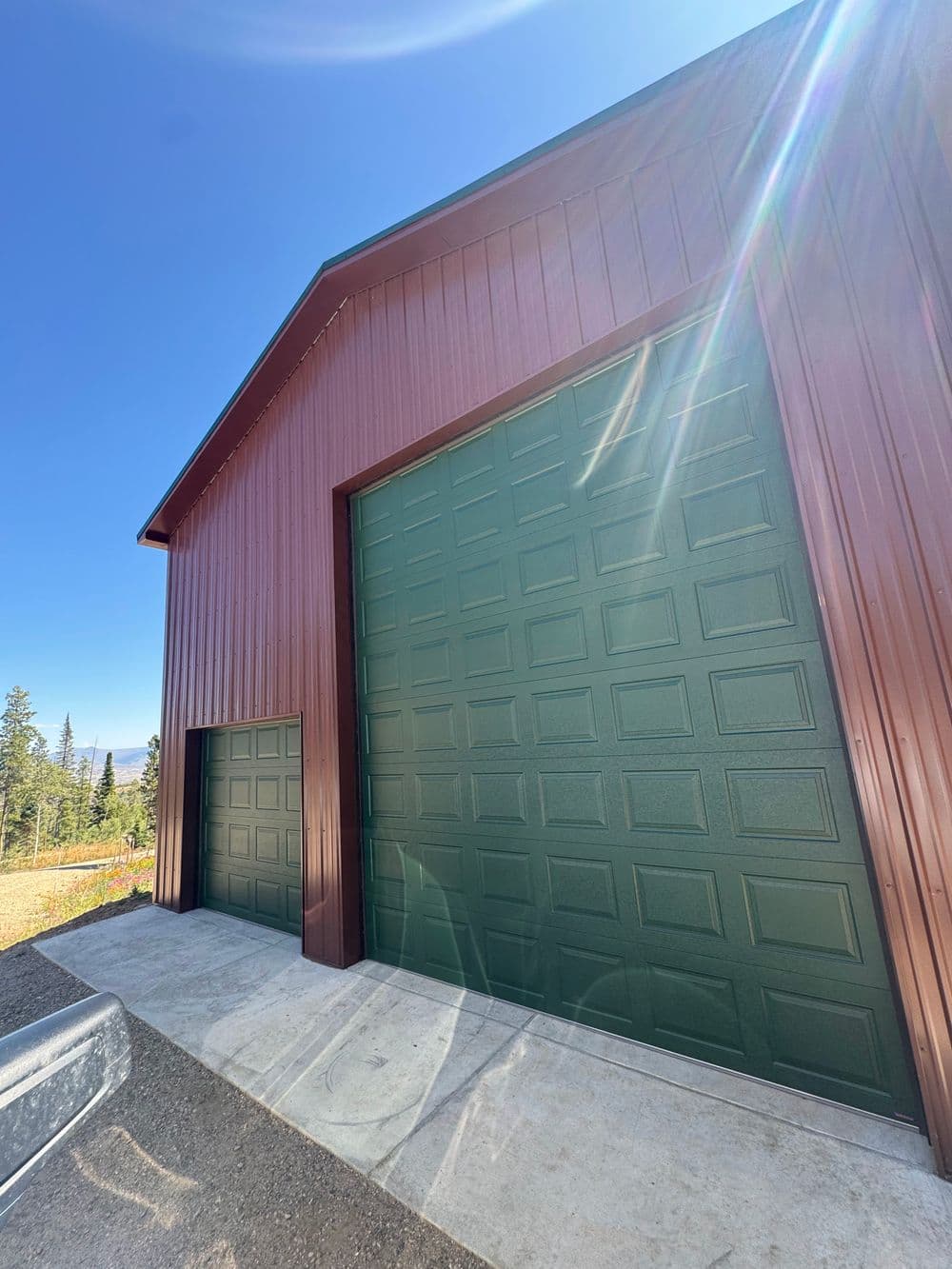 Red metal garage with two green doors against a clear blue sky and trees in the background.
