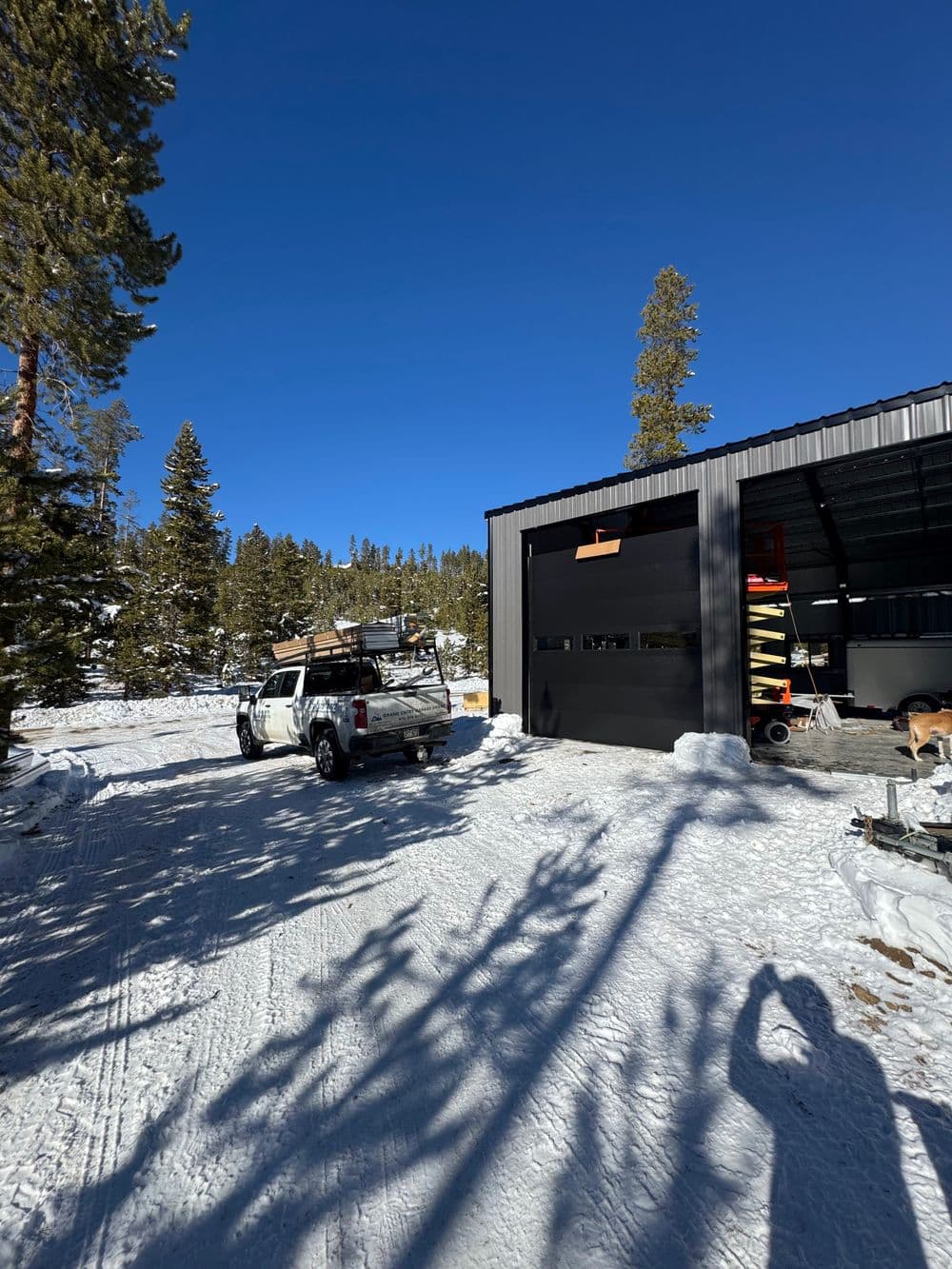 Snowy mountain landscape with a truck outside a metal garage and pine trees under a blue sky.