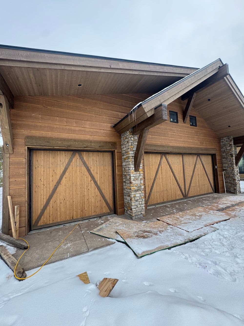 Modern wooden garage with two doors, stone accents, and snow-covered ground.