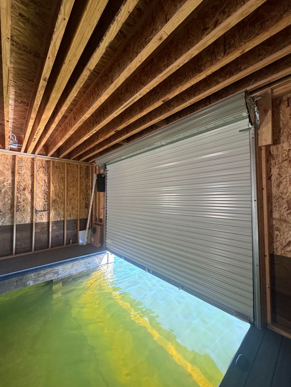 Rolling garage door opening to reveal a pool area with green water inside a wooden structure.