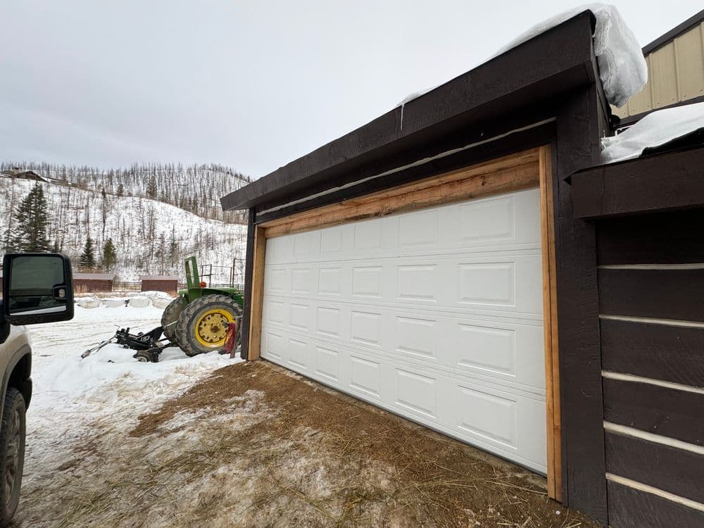 Tractor beside a white garage door in a snow-covered rural area with mountains in the background.