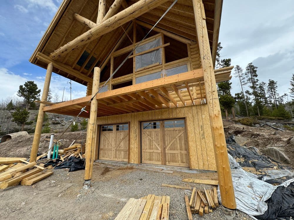 Modern wooden cabin under construction with a spacious deck and garage doors surrounded by nature.