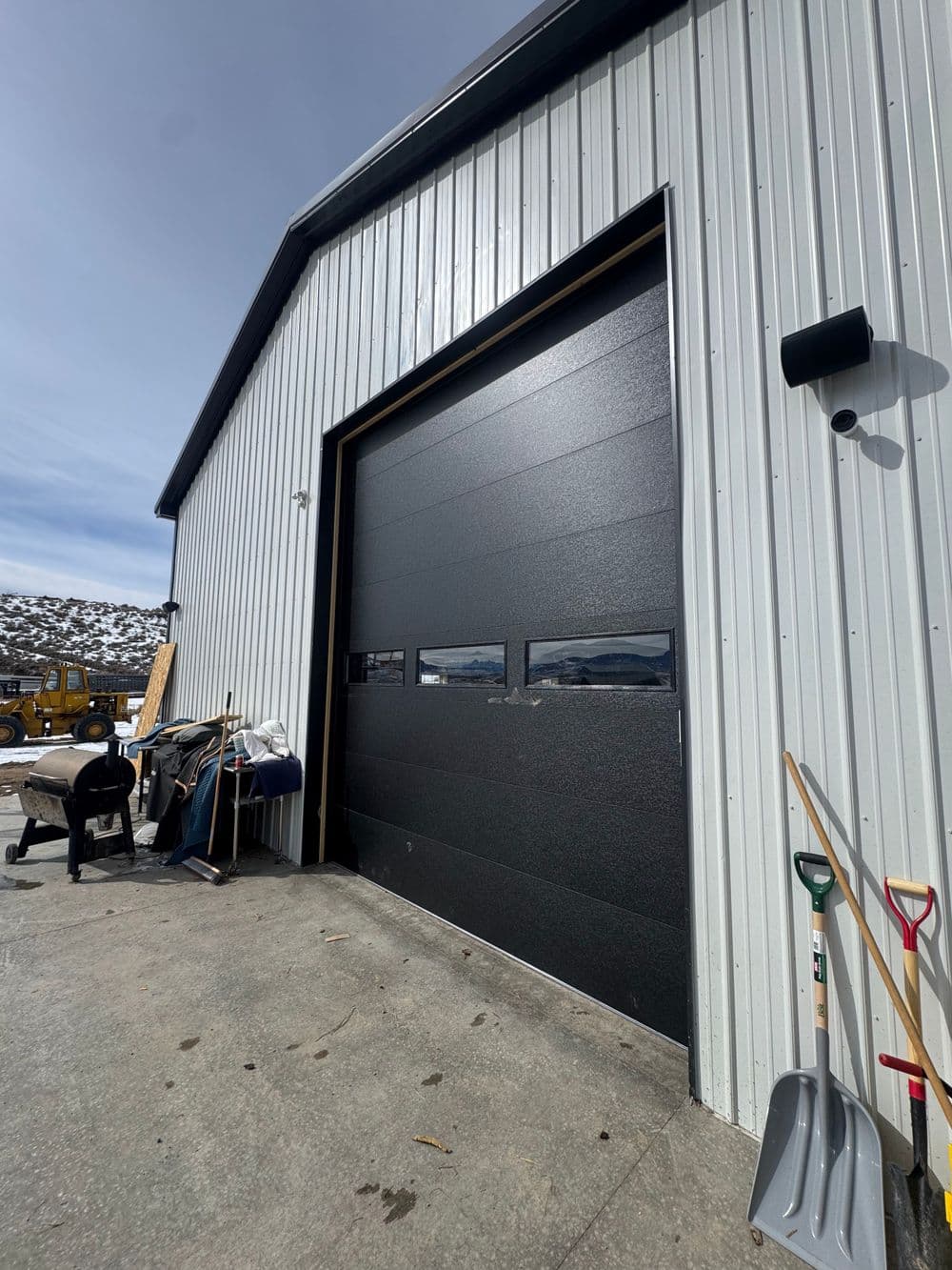 Metal storage building with large black garage door and tools outside. Snowy landscape in background.