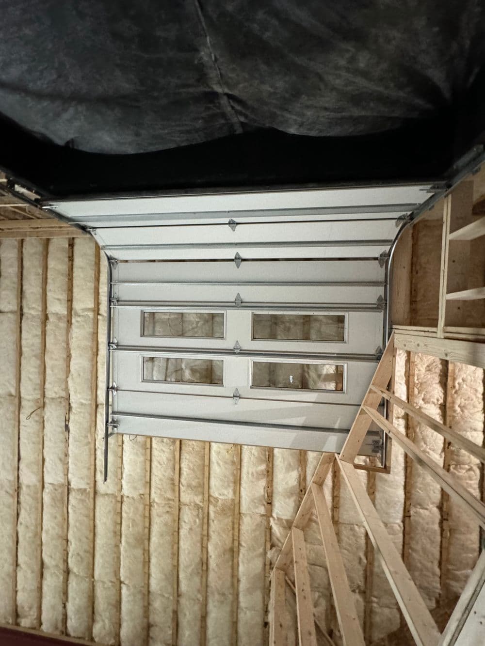 Insulated garage door with windows, framed by wooden structure and exposed wall insulation.