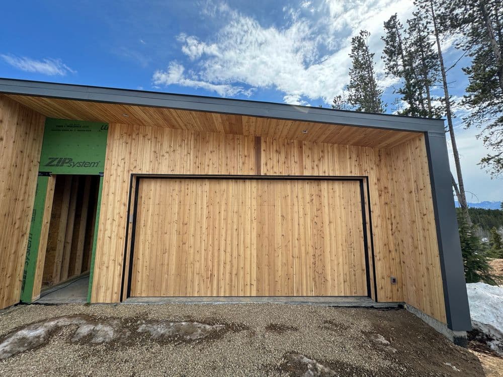 Modern wooden garage door with natural finish in a forested area under a blue sky.