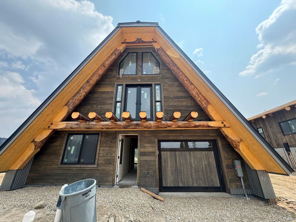 Modern A-frame house with wooden beams, large windows, and a gravel foundation under a blue sky.