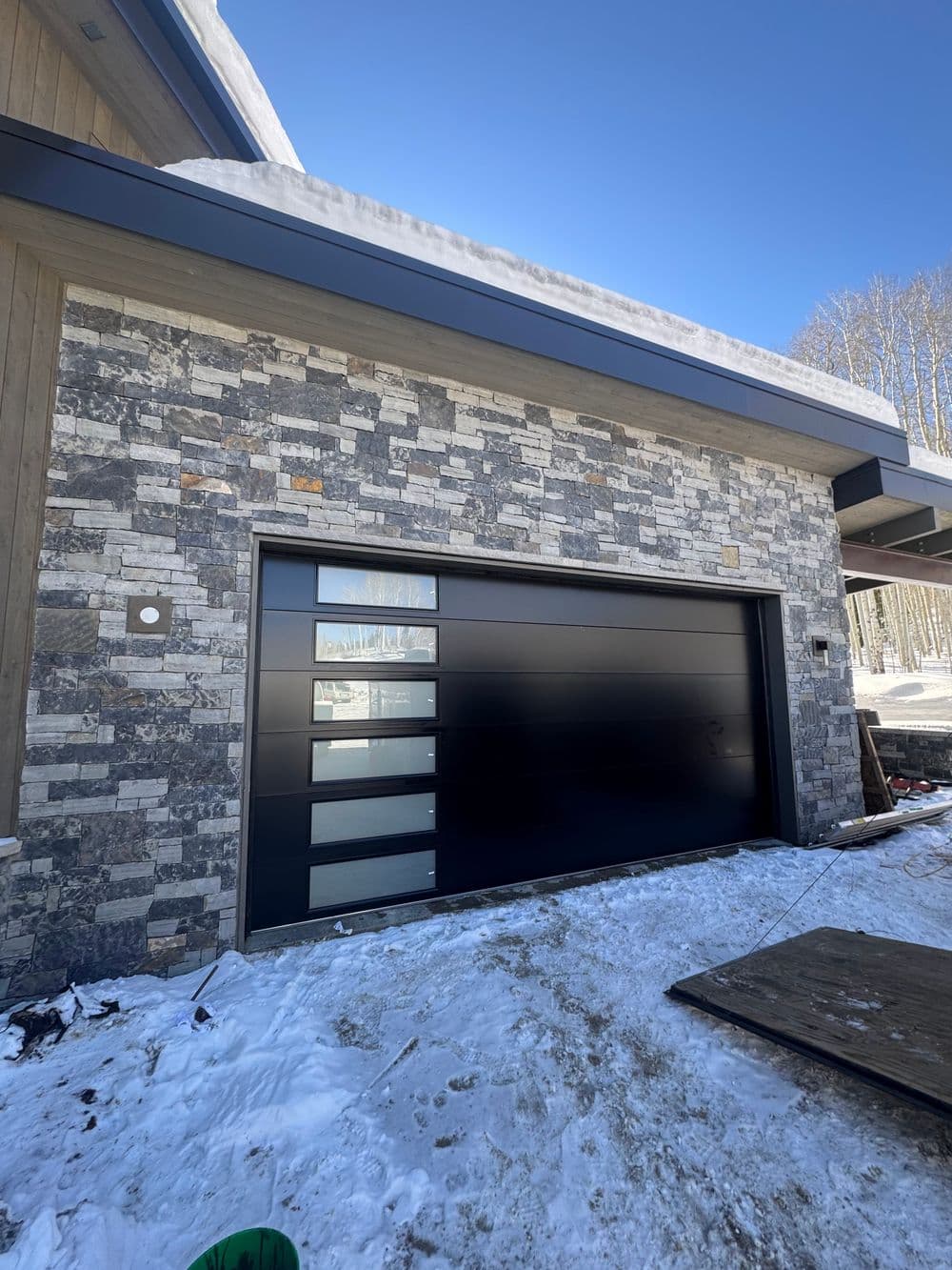 Modern black garage door on a stone-clad home exterior in winter setting with snow.