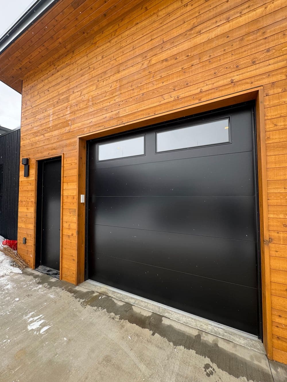 Modern black garage door and entrance door on a wooden exterior home. Snowy driveway.