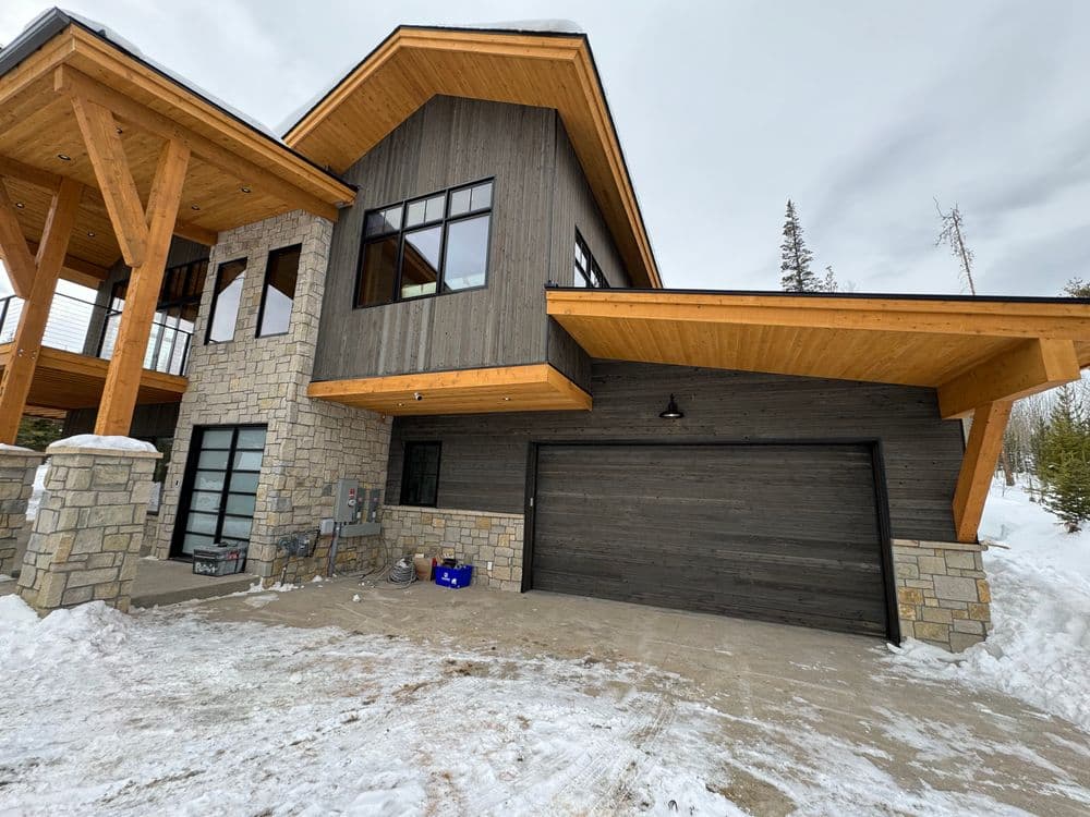 Modern mountain house with stone and wood exterior, garage, and snowy surroundings.