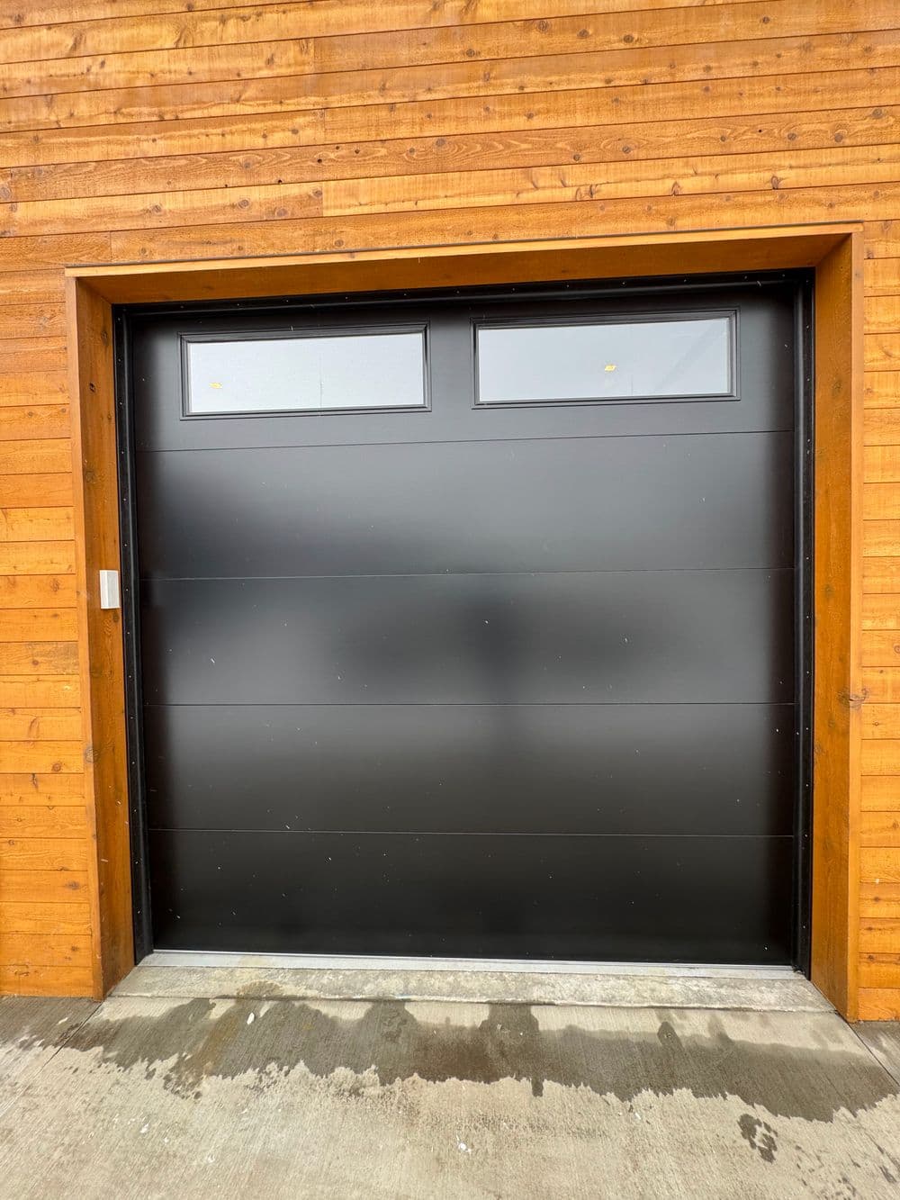 Modern black garage door with upper windows against a wooden panel background.