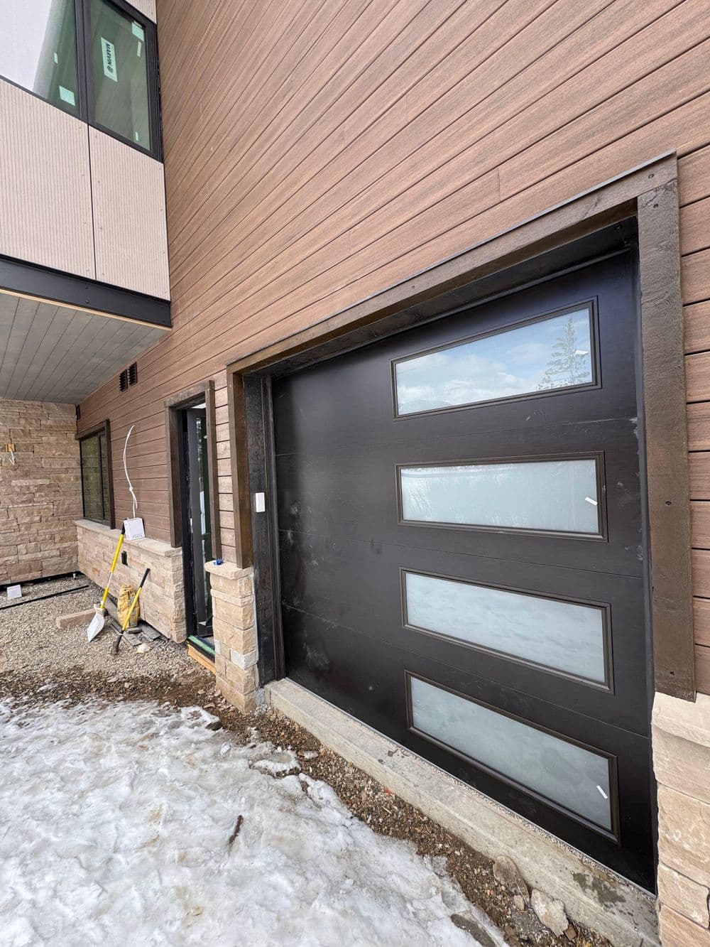 Modern black garage door with large windows, next to a contemporary home under construction.