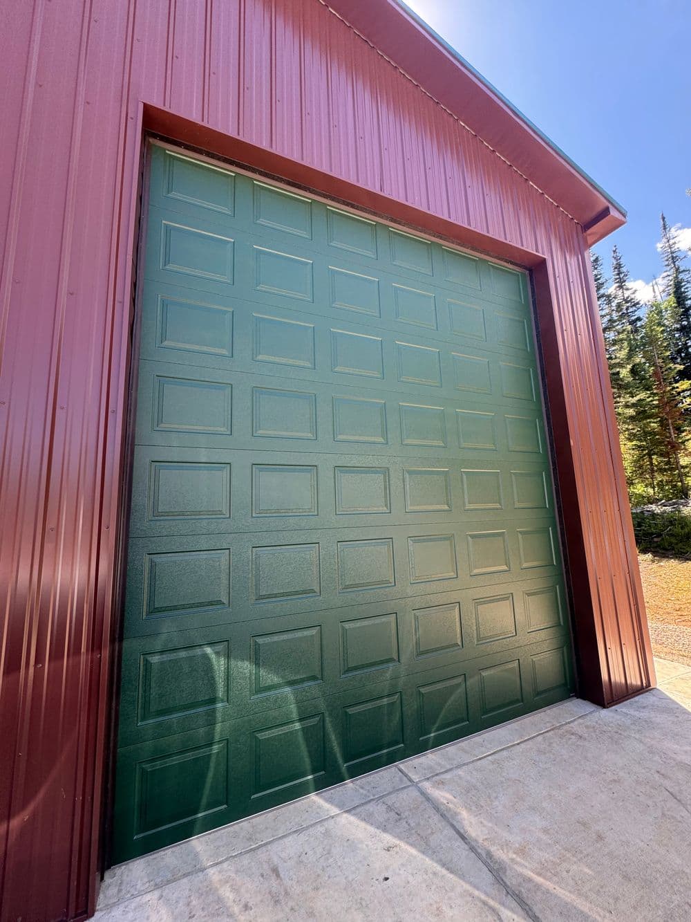 Green garage door on a red metal building with a clear blue sky and trees in the background.