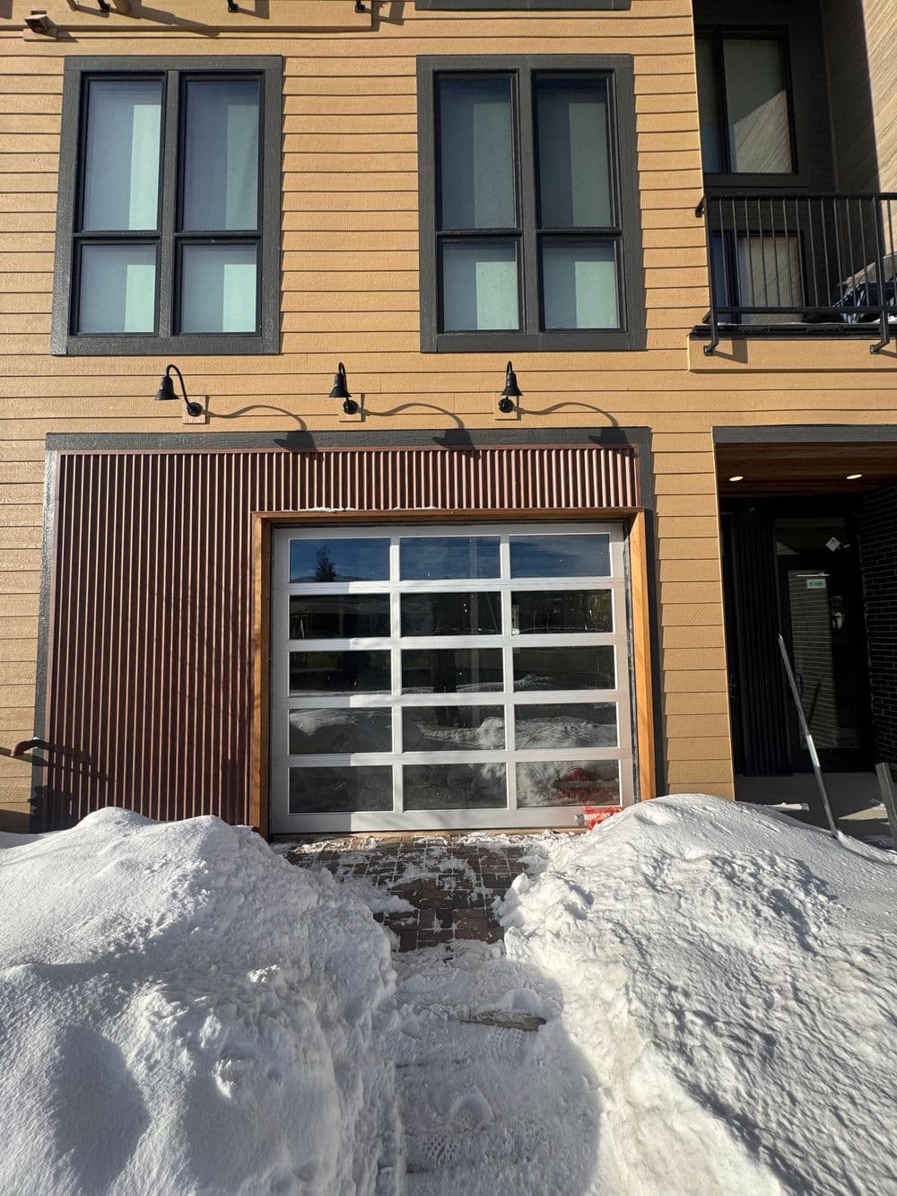 Modern home exterior with a glass garage door and snow-covered walkway. Natural light filters in.