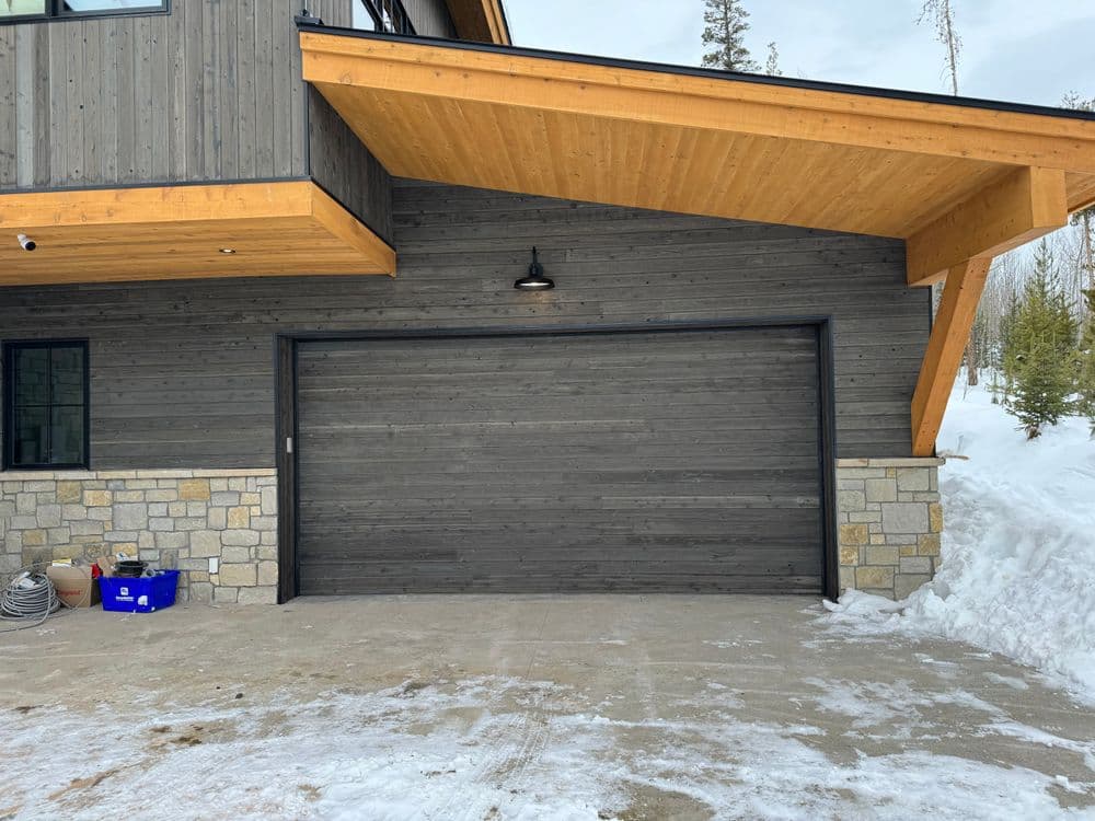 Modern home exterior featuring a dark wooden garage door and stone accents under a wooden overhang.