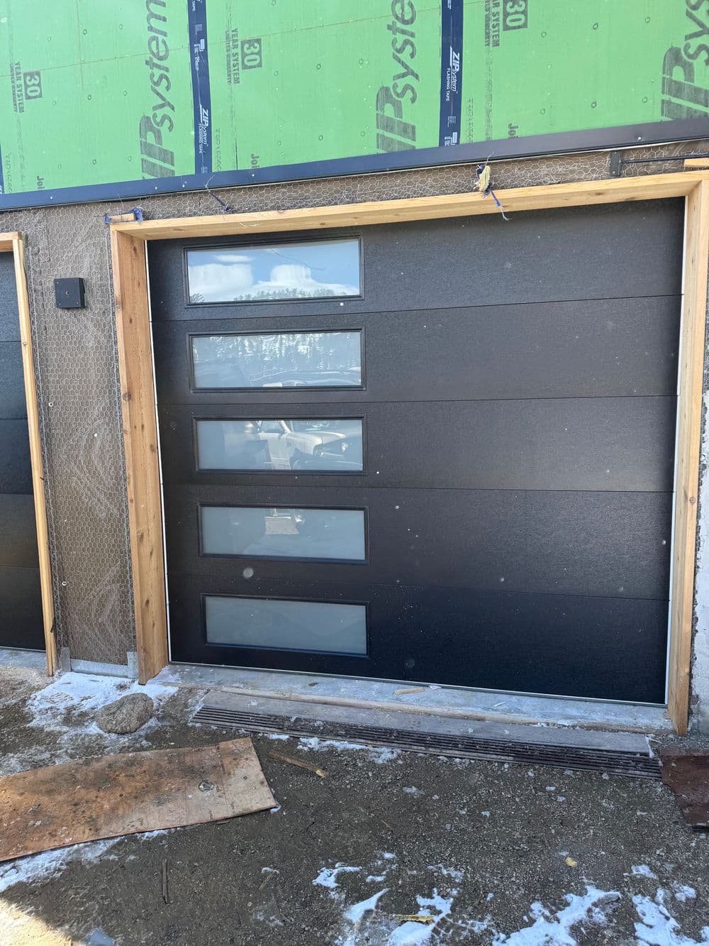 Modern black garage door with four inset glass panels, set against construction background.