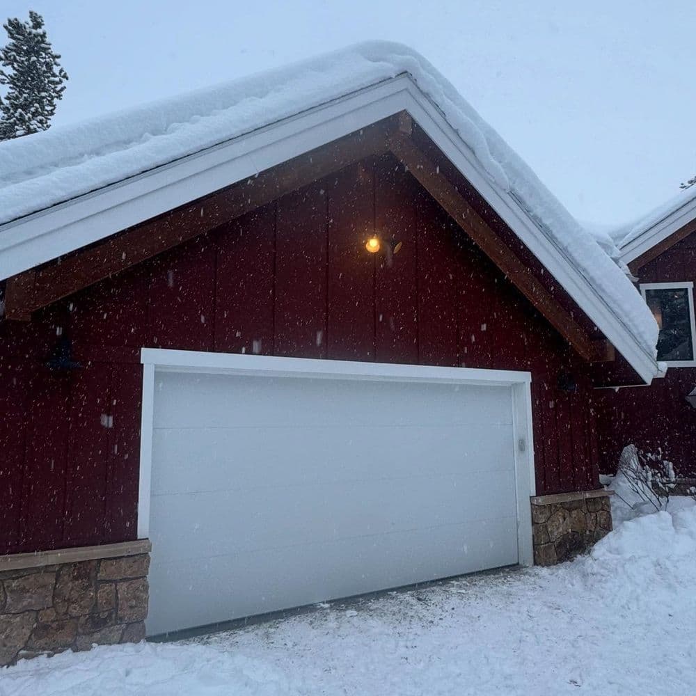 Snow-covered red cabin with a white garage door under a snowy evening sky.