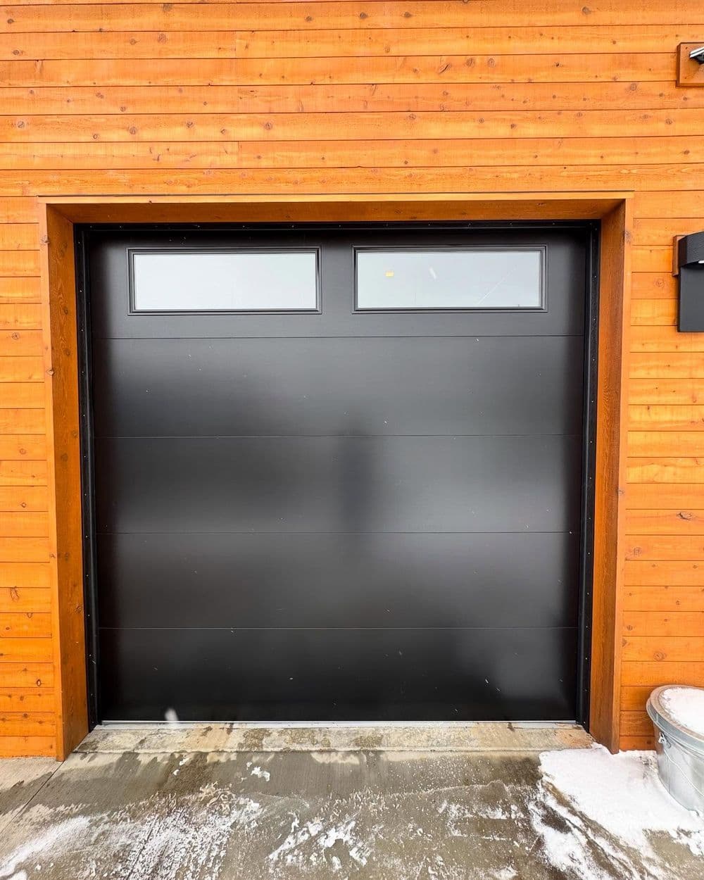 Modern black garage door with windows, set against a wooden exterior wall.