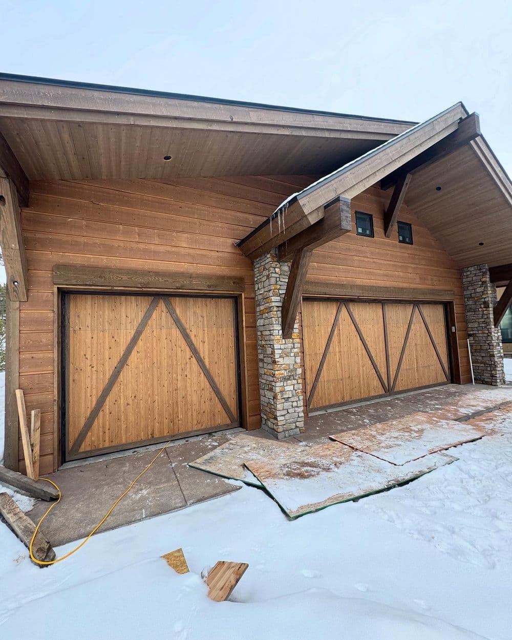 Modern wooden garage with stone accents, snowy landscape, and construction materials nearby.
