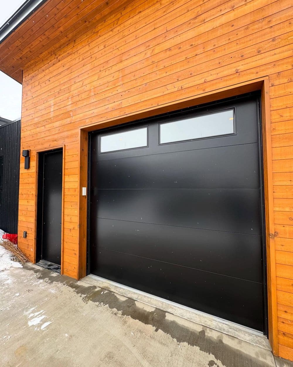 Modern black garage door on a wooden cabin exterior with a side entrance. Snow on the ground.