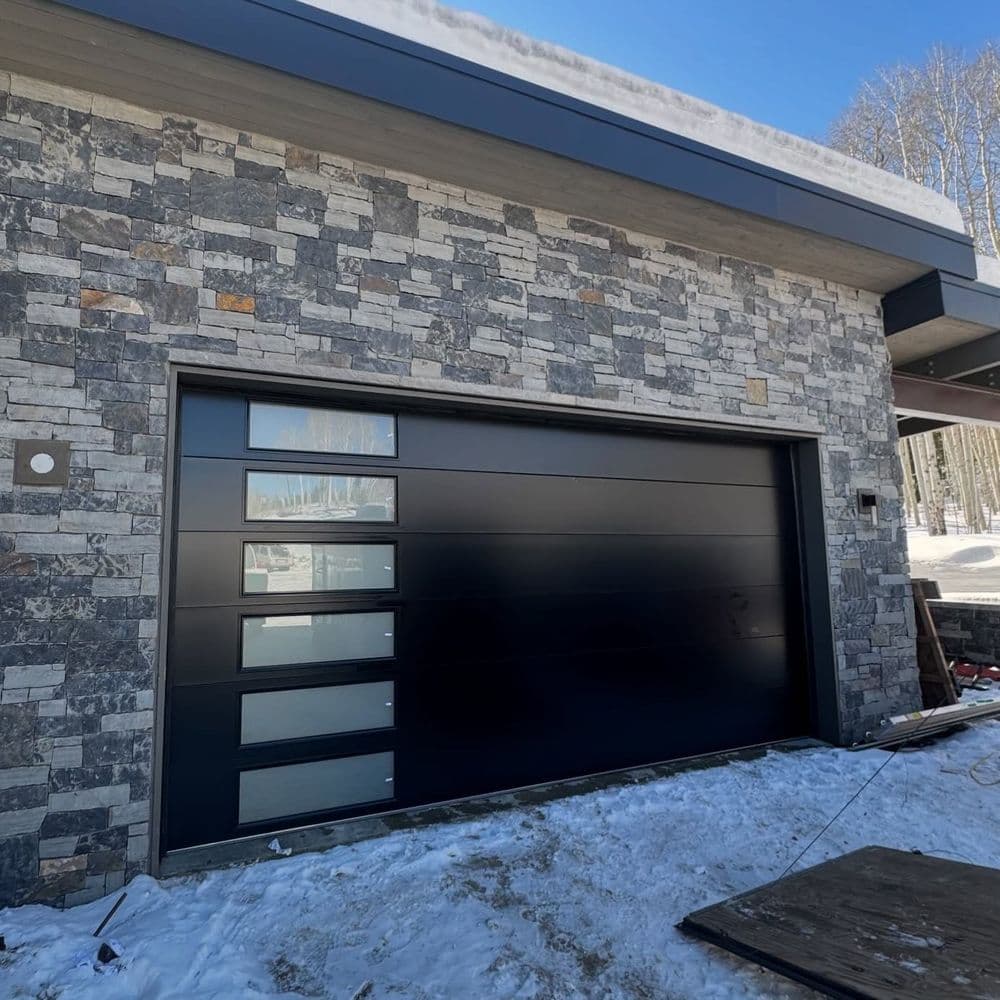 Modern black garage door on stone wall, surrounded by snow-covered landscape.
