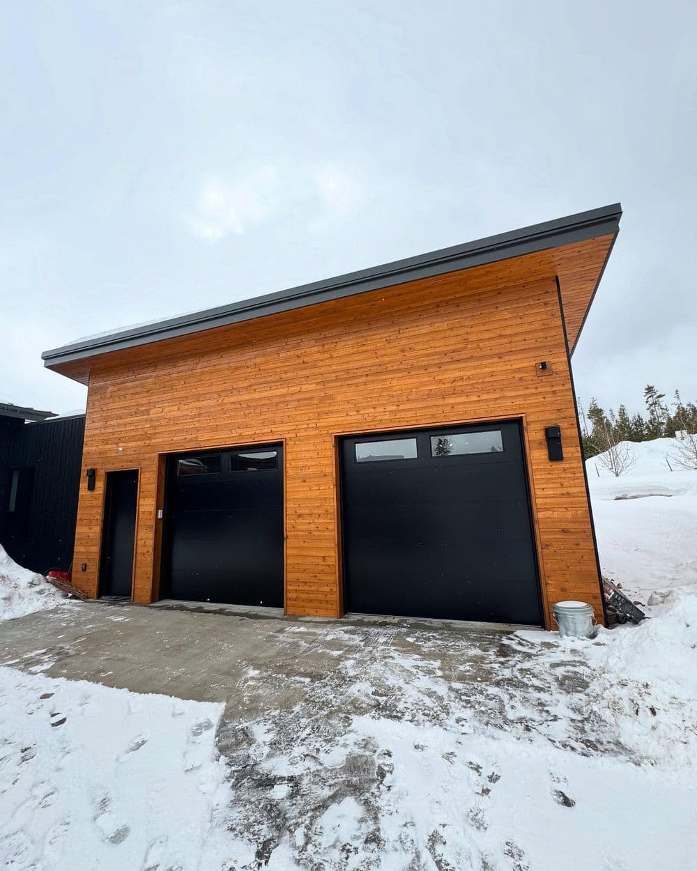 Modern wooden garage with two black doors, set in a snowy landscape.